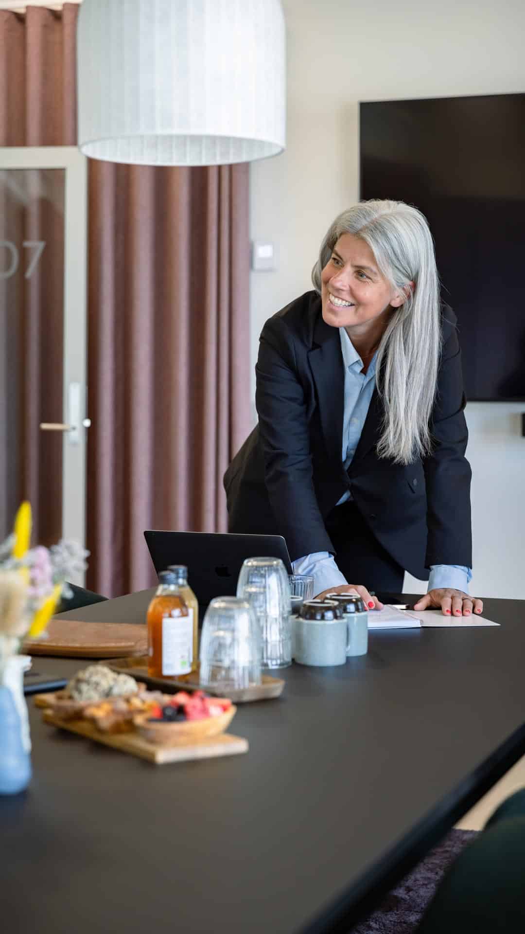 Business kvinde gør med et smil klar til præsentation i et mødelokale med stor tv-skærm. På bordet står forplejning i form af vand, juice og sunde snacks klar